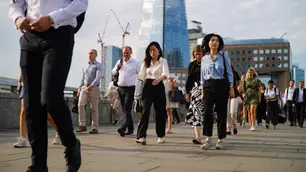 People walk on a busy city street with skyscrapers and cranes in the background. The mood is bustling under a partly cloudy sky.