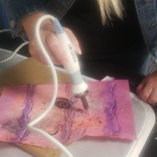 A woman holding a craft soldering iron making decorative holes in a piece of Lutradur that has had surface design applied