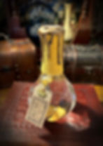 The Elixir of Midas Touch Potion. A larger potion with a classic potion bottle shape. Inside is a clear liquid. The neck is wrapped in yellow cord, topped with drippling metallic golden wax and topped with a silver seal. A brown label hangs from the neck with a golden crown charm attached.