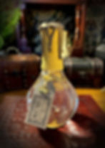 The Elixir of Midas Touch Potion. A larger potion with a classic potion bottle shape. Inside is a clear liquid. The neck is wrapped in yellow cord, topped with drippling metallic golden wax and topped with a silver seal. A brown label hangs from the neck with a golden crown charm attached.