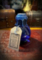 The Potion of Mana. A small, rounded glass bottle, contains a deep blue liquid. The neck is wrapped in drak blue cord and topped with a metallic blue wax, topped with a silver seal. A brown label hangs from its neck with a small silver moon charm attached.