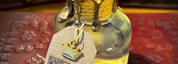 The Elixir of Midas Touch Potion. This small ink well shapped glass bottle is full of clear liquid. Its neck is wrapped in a deep yellow cord, topped with dripped metallic gold wax and topped with a silver seal. Hanging from its neck is a brown label adorned with a small gold crown charm.