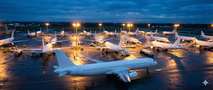 A high-angle view of a busy aerospace delivery center at twilight, crowded with multiple unbranded, plain white Airbus A320neo and A321neo aircraft. Ground crews and support vehicles surround the jets under bright floodlights, while one white aircraft taxies for departure in the foreground against a deep blue evening sky.