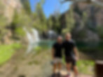 Two men pose on a boardwalk in front of a multi-tiered waterfall and clear lake.