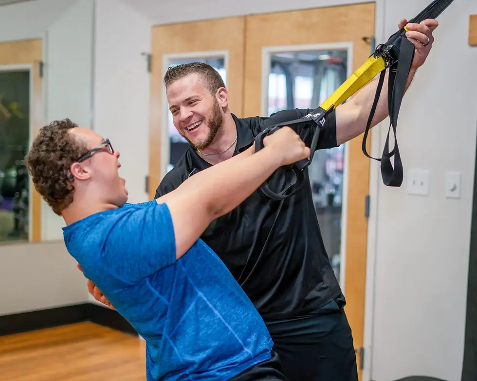 Man assisted by trainer using suspension straps, both smiling.