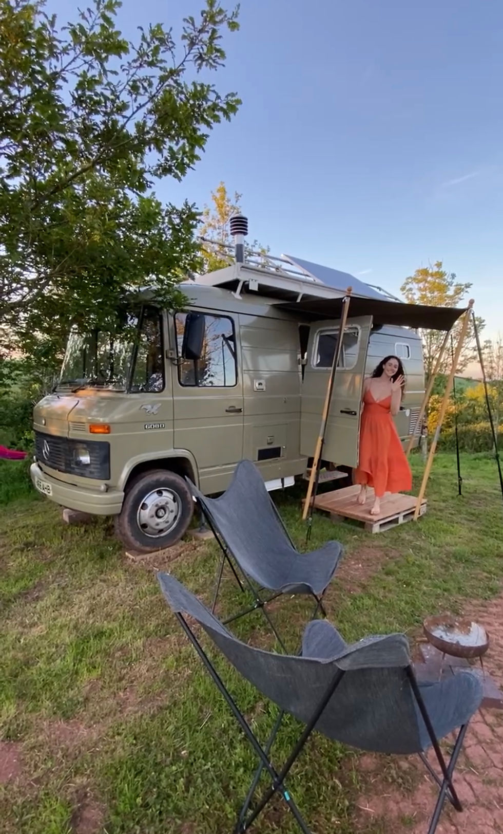 A woman stands outside a khaki van with a front porch, solar panel on top, and chairs out front.