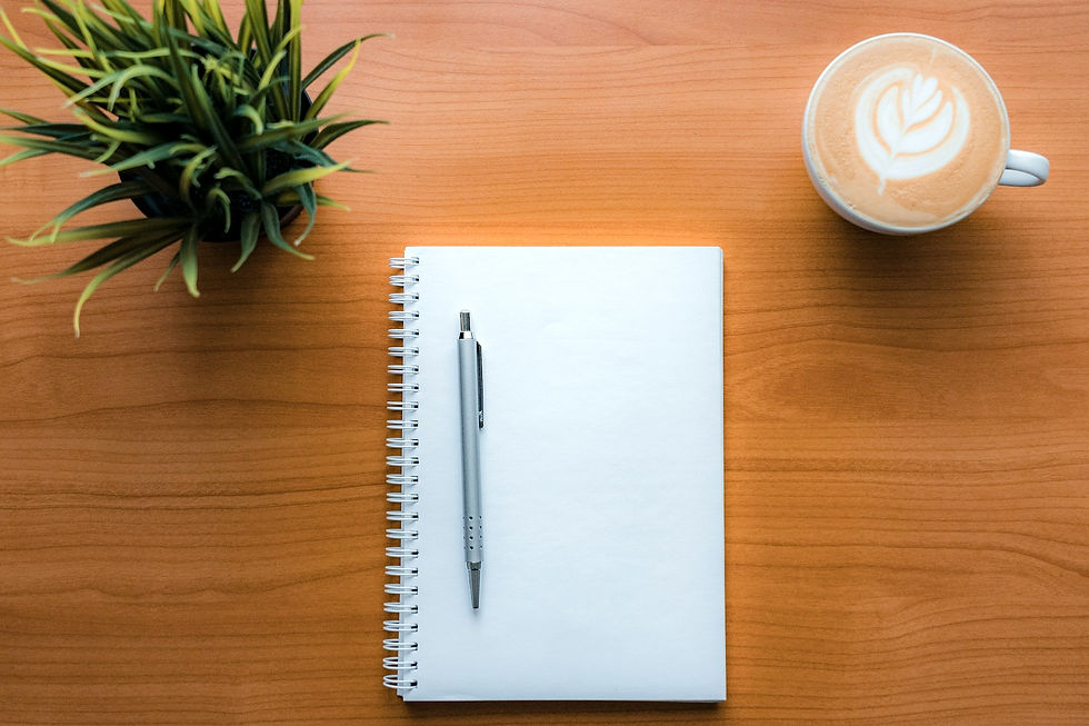 Notepad with pen on a wooden table, latte art coffee, and a potted plant create a calm workspace setting.