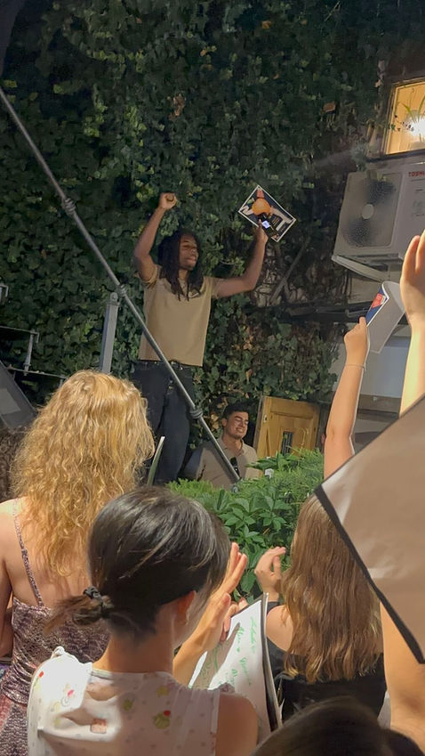 Against an ivy-covered wall at night, a person raises both arms in celebration while a boom pole and a hand holding a small card appear in the foreground.