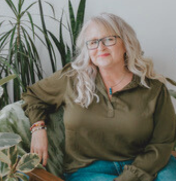 Reiki Master Cara Holmstrup seated in a peaceful studio with plants, wearing olive top and jeans