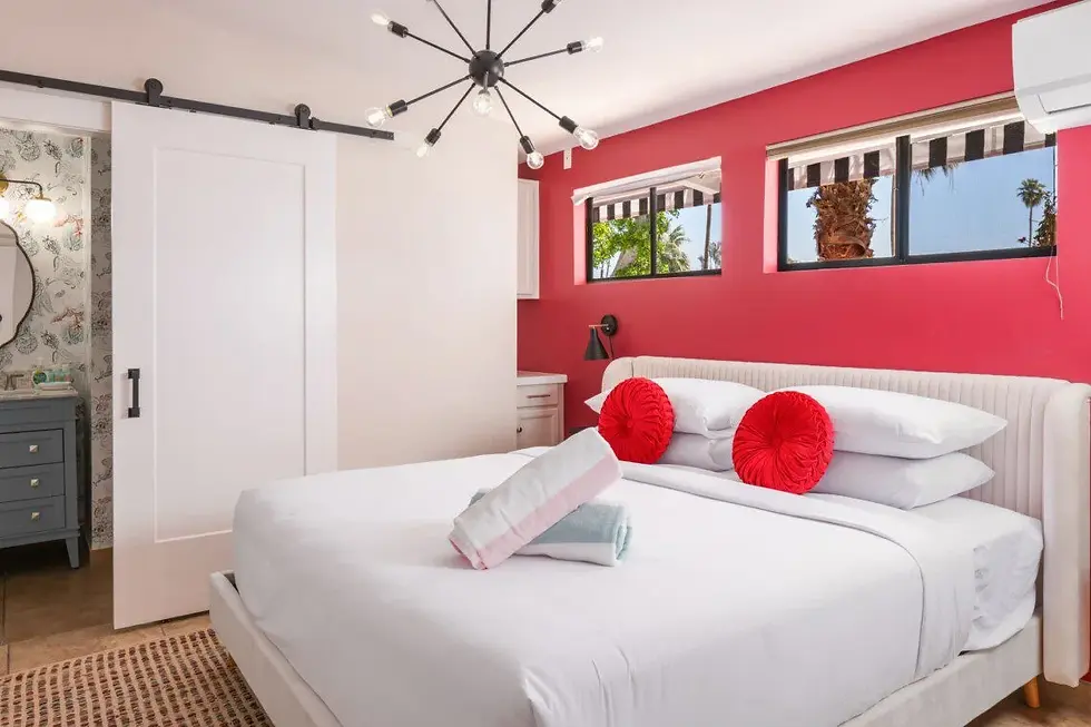 Modern Palm Springs hotel bedroom with coral pink accent wall, queen bed with red pillows, and windows overlooking palm trees