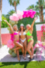 Three women in bikinis smiling and posing by a colorful balloon arch outdoors. Palm trees and a festive table are visible in the sunny setting.