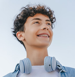 Junge mit fester Zahnspange und Kopfhörern lächelt vor blauem Himmel. 