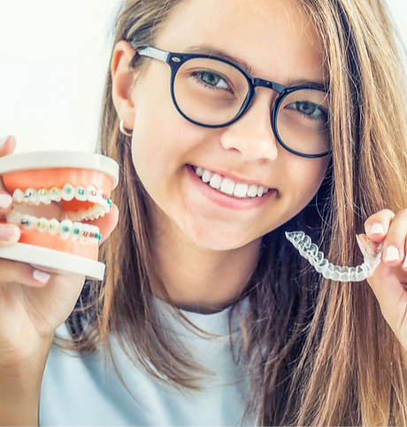 Jugendliches Mädchen mit Brille hält in der einen Hand ein Zahngebiss mit bunter Zahnspange und in der anderen Hand hält sie eine lose Zahnspange ins Bild