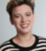 Smiling person with short, two-toned hair wearing a black and white striped sweater, standing against a plain gray background.