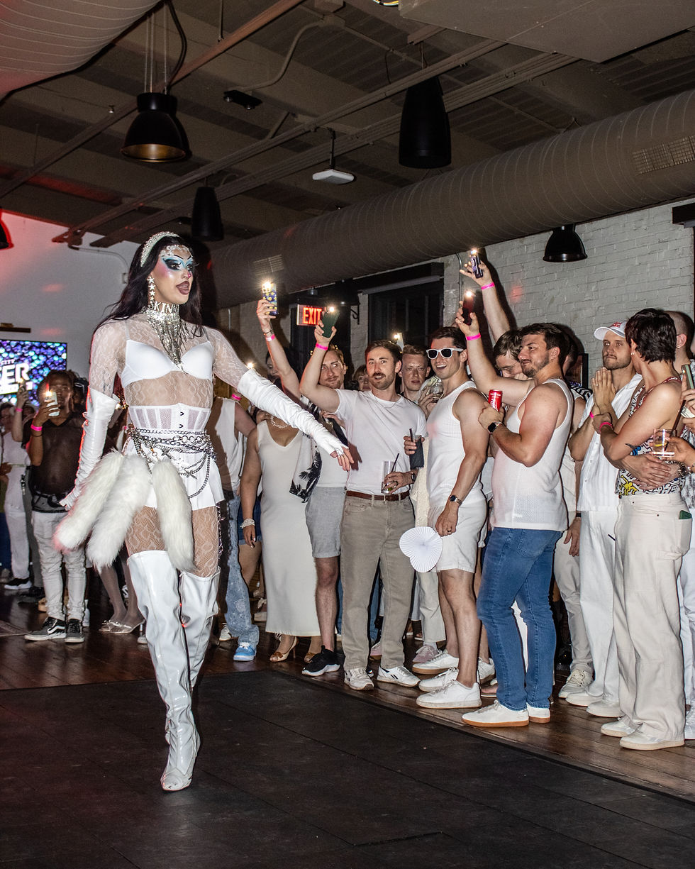 Drag performer in white lace and boots struts past cheering crowd holding lit phones in a club, with an EXIT sign in back.