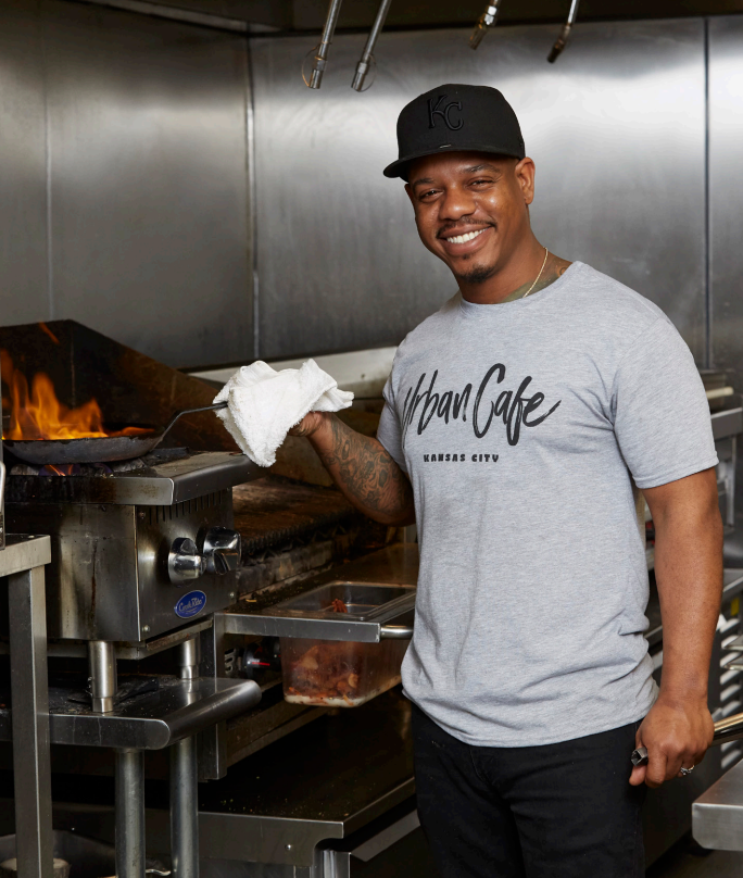 Smiling cook in a gray "Urban Cafe Kansas City" shirt holds a skillet by a stove with flames. Industrial kitchen setting, black cap.