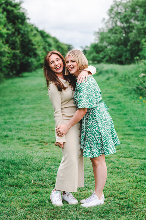 Two sisters hugging and laughing together in a lush green meadow during a relaxed outdoor family portrait session in Wiltshire