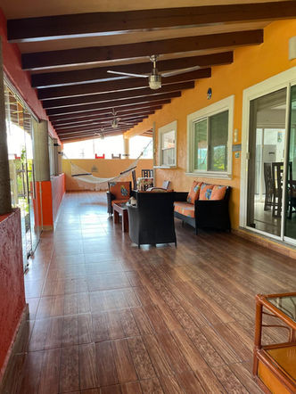 Amplia terraza techada con techo de vigas de madera y ventiladores, decorada en tonos cálidos anaranjados y rojizos, ideal para disfrutar al aire libre. Terraza con muebles de mimbre y cojines decorativos, hamaca colgada para relajación y ventanales de vidrio corredizos que conectan al interior de la casa. Piso de cerámica estilo madera que brinda un toque acogedor. Espacio perfecto para reuniones familiares o para disfrutar de una tarde tranquila en casa.