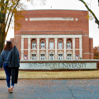 Winthrop University Graduate walking in front of campus sign with gown over shoulder 