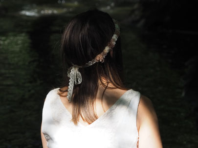 Silhouette féminine en robe blanche et couronne florale, baignée de lumière dans l’ombre d’un sous-bois.