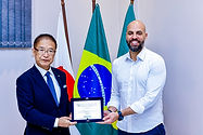 A Prefeitura de Osasco realizou segunda-feira, 3/8, no Paço Municipal, evento em comemoração aos 50 anos de irmanamento entre o município e a cidade de Tsu, no Japão. O encontro reuniu a delegação japonesa, autoridades municipais, representantes da comunidade nipo-brasileira e sociedade civil.
O acordo de cidades-irmãs, também conhecido como irmanamento, é uma parceria firmada entre municípios de diferentes países com o objetivo de fortalecer os laços de amizade, promover o intercâmbio cultural, educacional, econômico e institucional, além de incentivar a cooperação em áreas de comum interesse. A relação entre Osasco e a cidade japonesa de Tsu teve início em 1976, quando foi firmado o primeiro acordo de irmanamento entre os dois municípios.
A programação teve início com a recepção oficial da comitiva japonesa. O prefeito Gerson Pessoa deu as boas-vindas à delegação de Tsu, composta pelo prefeito Yasuyuki Maeba, pelo presidente da Câmara Municipal, Kin’ichi Ono, pelo secretário-geral, Katsutoshi Ochiai, pela vice-diretora de Intercâmbio e Relações Públicas, Naomi Yamaguchi, pelo presidente Hisahiko Yamaguchi, pelo consultor Nobuo Sato, pelo diretor de Recursos Humanos e Assuntos Gerais, Yukinori Yamada, pelo CEO presidente e diretor representante Tomokazu Hayashiguchi e pelo consultor sênior Tetsuo Yoshimura. "É uma grande alegria receber a delegação da cidade de Tsu em Osasco. Neste ano em que celebramos os 50 anos do nosso acordo de cidades-irmãs, reafirmamos o compromisso de fortalecer essa parceria por meio da troca de experiências, conhecimentos e culturas entre os nossos municípios”, disse Gerson.
Em seguida, as autoridades participaram da assinatura do termo de reafirmação do acordo de cidades-irmãs entre Osasco e Tsu. Na sequência, a comitiva visitou a Galeria dos Prefeitos, na recepção do Paço Municipal, acompanhada pelos ex-prefeitos de Osasco Silas Bortolosso e Francisco Rossi.
O prefeito de Tsu, Yasuyuki Maeba, destacou a emoção de participar da celebração. "É uma sensação maravilhosa. Apesar das 31 horas de viagem entre Tsu e Osasco, é muito gratificante estar aqui para celebrar este marco tão importante. Chegamos hoje pela manhã e, no mesmo dia, já tivemos a oportunidade de reafirmar oficialmente esse tratado de amizade, o que nos deixa muito felizes".
O prefeito japonês também ressaltou as expectativas para o futuro da parceria entre as duas cidades. "Desta vez trouxemos também empresários de Tsu para fortalecer essa parceria. Esperamos que, a partir deste novo ciclo, possamos ampliar o intercâmbio empresarial e criar novas oportunidades de negócios, construindo juntos os próximos 50 anos dessa relação entre Tsu e Osasco".
Para encerrar a programação, foi realizado o tradicional hasteamento das bandeiras. A de Osasco foi hasteada pelos vereadores presentes, a do Brasil por Gerson Pessoa, e a do Japão por integrantes da delegação japonesa, simbolizando os laços de amizade, cooperação e respeito entre as duas cidades-ir
