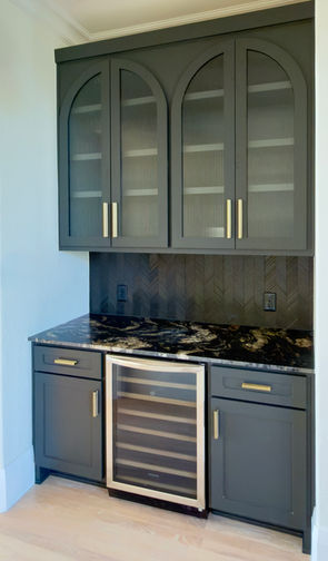 Built-in bar area between kitchen and dining room with black cabinetry, arched ribbed glass-front doors, textured glass tile backsplash, brushed brass hardware and wine refrigerator.