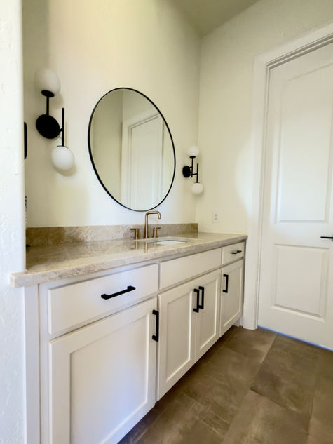 Modern bathroom vanity with creamy white shaker cabinetry, quartz countertop, round black-framed mirror, and matte black hardware, styled with warm neutral concrete look flooring.