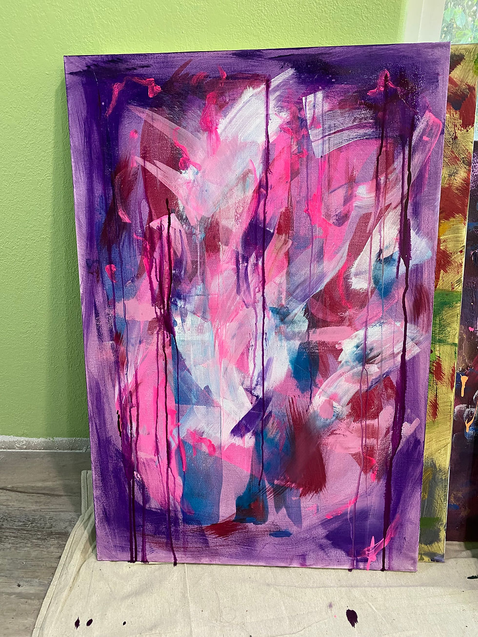 Abstract painting with vivid pink, purple, and white brushstrokes on canvas. Drips create a dynamic feel. Set against a light green wall.