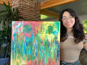 Woman smiling, holding a colorful abstract painting in a cozy room with plants and brick walls. She's wearing a striped shirt and glasses.