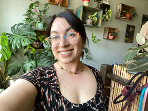 Smiling person with glasses takes a selfie in a room filled with lush green plants and wooden shelves. Patterned dress and warm ambiance.