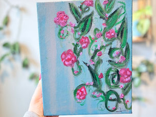 Hand holds a canvas with pink and green floral painting on blue background. Blurred indoor plants visible in the soft-focus background.