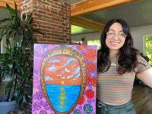 Woman smiling, holding a colorful painting of a sunset over water framed by roses. Indoor setting with brick and wood accents, dog nearby.