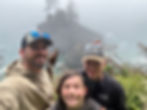 Three smiling hikers take a selfie on a foggy cliff above a rocky sea stack with trees; ocean waves below.