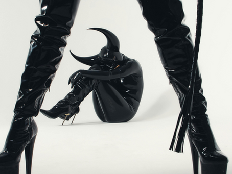 Foto di due persone vestite in latex: una è accucciata a terra, l'altra la sovrasta.