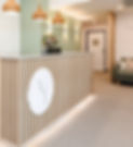 The Nourish & Thrive reception area, with light timber reception desk and white Nourish & Thrive icon, overhead timber look pendant lights, soft wall lighting, a bunch of pink flowers and green foliage in a vase, a sage green lounge with cushions, and a consultation room door in the background.
