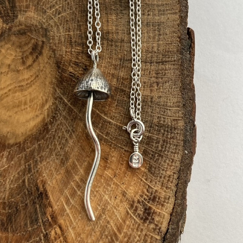 Sterling silver magic mushroom pendant displayed on a wood slice. The chain shows a bolt ring clasp with a stamped 925 tag.