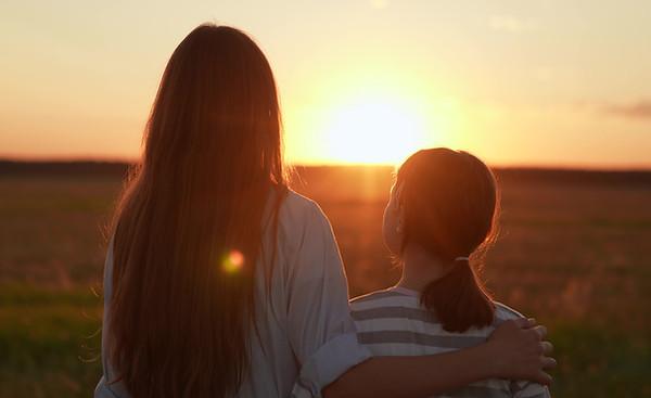 Eine Frau und ein Kind von hinten betrachtet blicken gemeinsam in einen warmen Sonnenuntergang; Symbolbild für die Arbeit mit dem Inneren Kind im IFS-Modell.