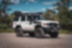 White off-road 76 Series LandCruiser on gravel, forest background, clear sky. Upgraded clutch, suspension, and roof rack. Adventure vibe.