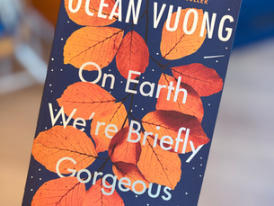 Hand holding Ocean Vuong’s On Earth We’re Briefly Gorgeous book cover with orange leaves on a dark blue background.