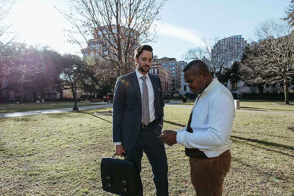 Two men in a park, one in a suit holding a briefcase and the other in casual wear checking a phone. Sunlight filters through trees.