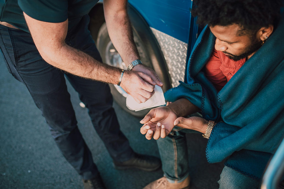 Man in blue blanket with injured hand gets care from another person on a sunny day, sitting by a blue vehicle. The mood is attentive.