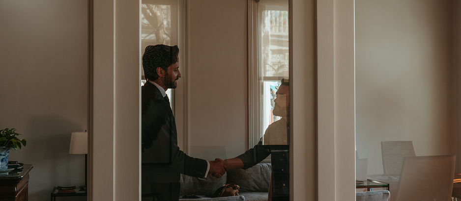 Two people shake hands in an office with a sofa and plants. The reflection on the glass door suggests a formal, professional mood.
