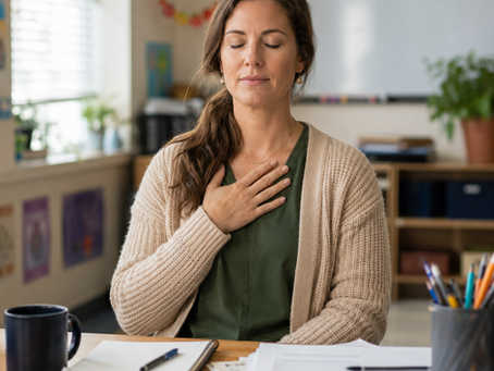 A teacher sits at her classroom desk with papers, notebooks, and pencils nearby, eyes closed and one hand on her chest, practicing a quiet mindfulness reset during the school day.