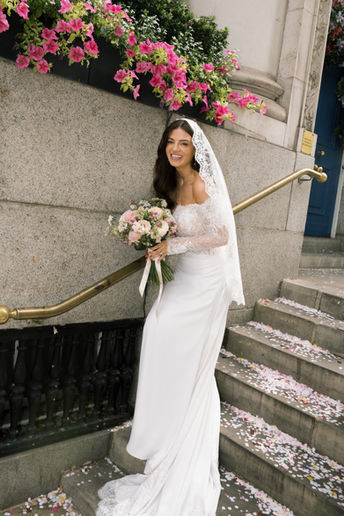 Smiling bride in a lace-trimmed veil holds a bouquet of pink and white flowers while standing on confetti-covered steps, beautifully captured by professional London wedding photographer Keidi Vaikma.