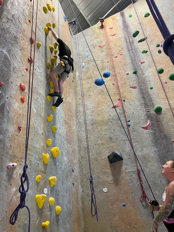 Two individuals at an indoor rock climbing gym, one is on the climbing wall and the other is holding the harness on the ground