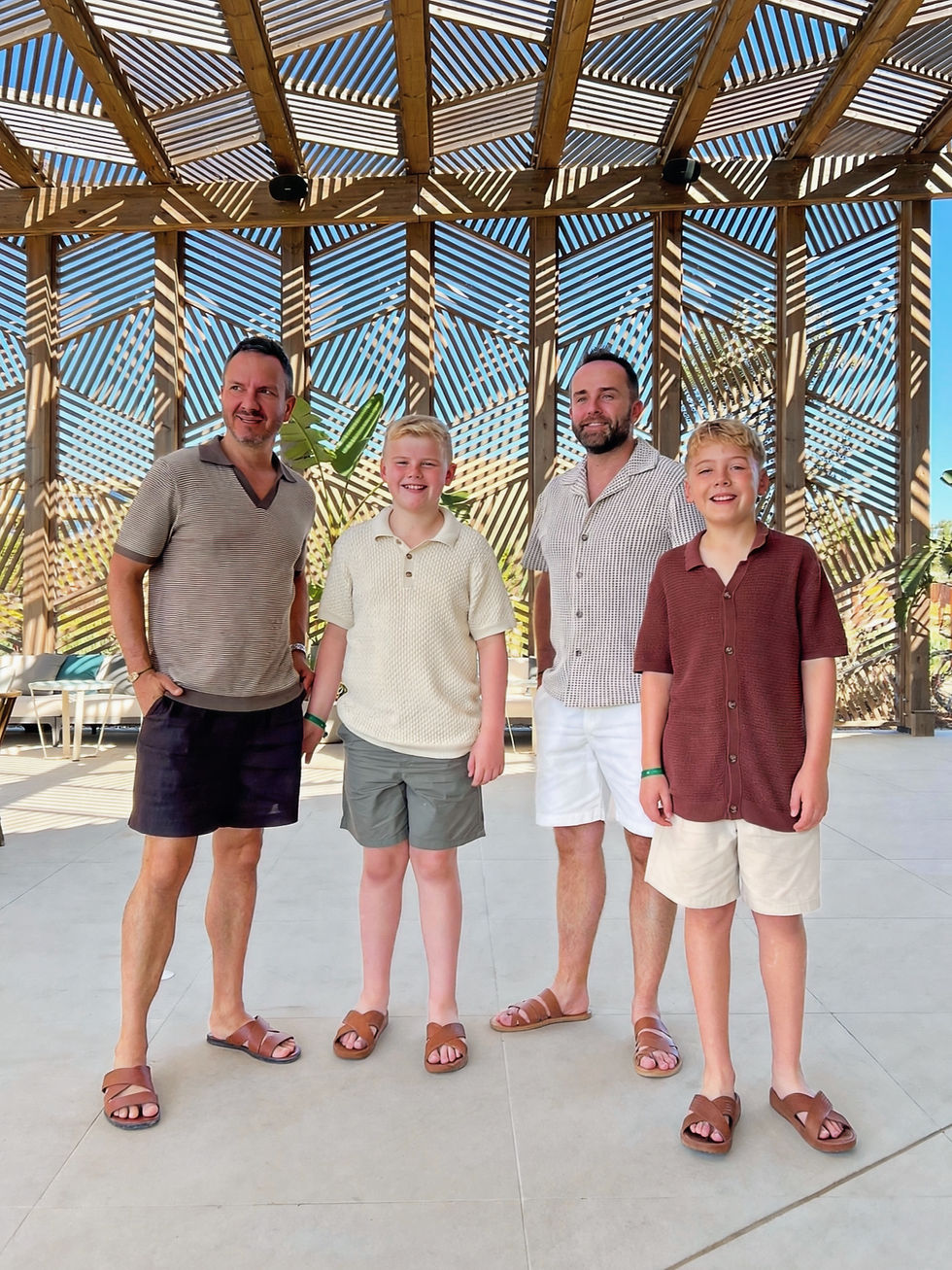 Four smiling males pose under a wooden pergola casting striped shadows, in shorts and sandals beneath a bright blue sky.