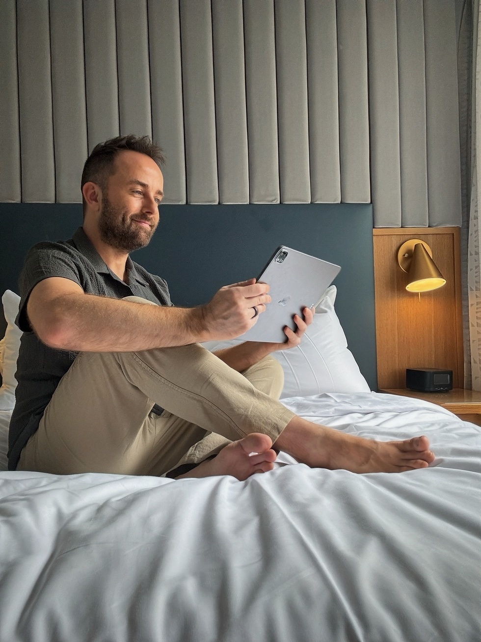 Man in a gray shirt smiles while using a tablet on a bed. Gray padded wall and a lit lamp in the background. Cozy, relaxed mood.