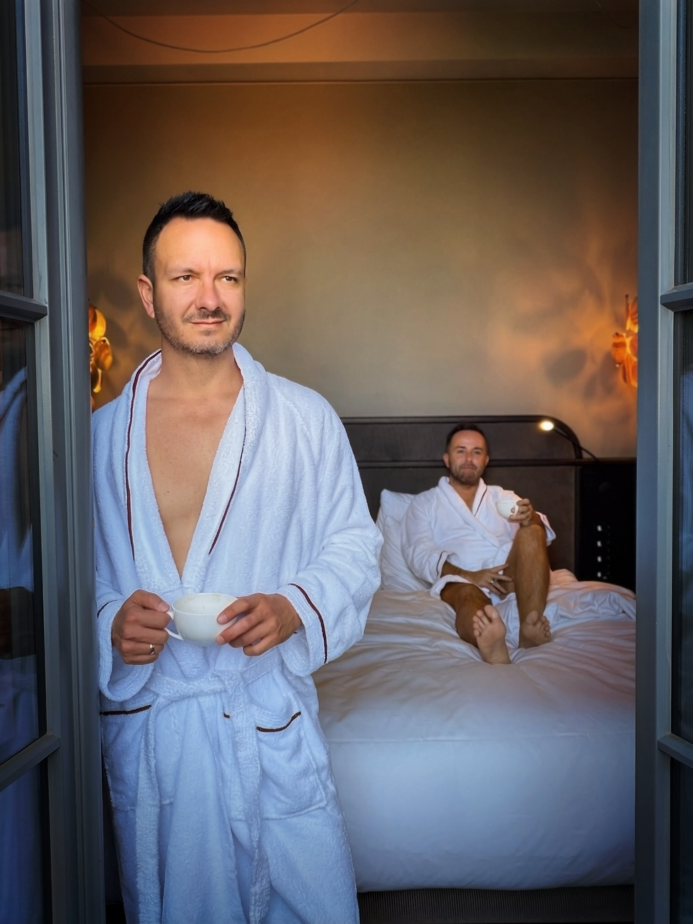 Male couple in robes in hotel room. One is standing with a coffee cup in hand while other is laying on bed