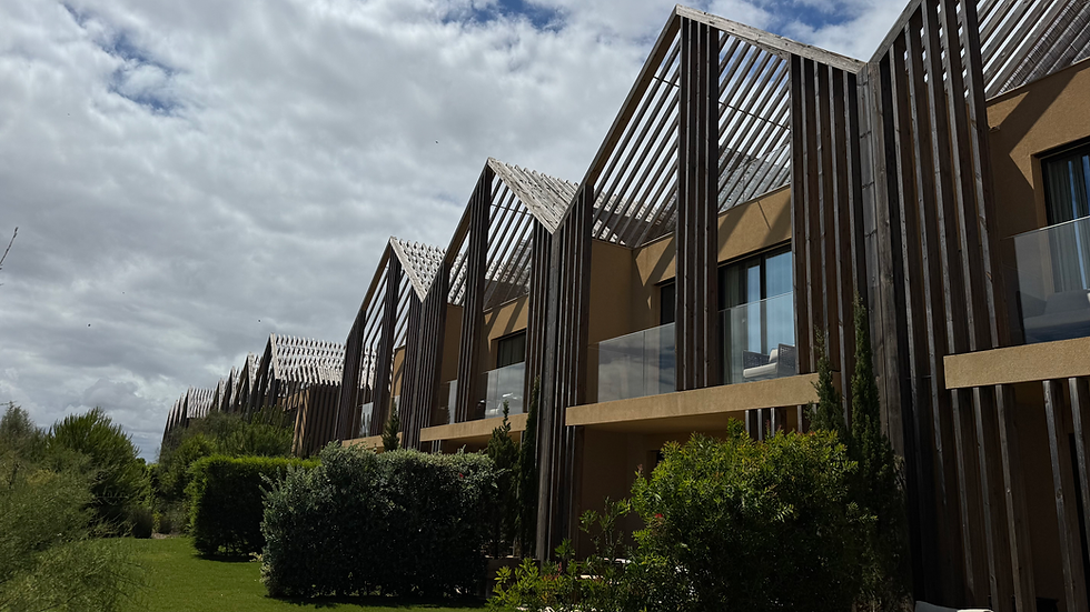 Modern resort-style row of tan buildings with wooden slatted roofs, glass balconies, and green shrubs under a cloudy sky