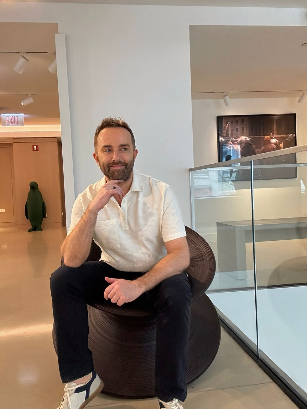 Man sitting thoughtfully on a modern chair in a gallery. White shirt, black pants. Art on walls, glass display. Warm, contemplative mood.